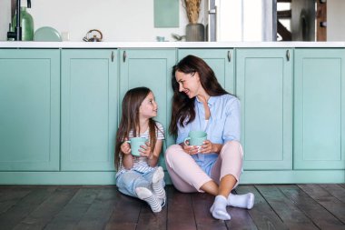 Gülümseyen mutlu anne ve kız ellerinde çay fincanları yemek odasında ahşap zeminde oturup komik hikayeler anlatıyor. Konforlu evde kahvaltı ediyorum. Hafta sonunu birlikte geçirmek.