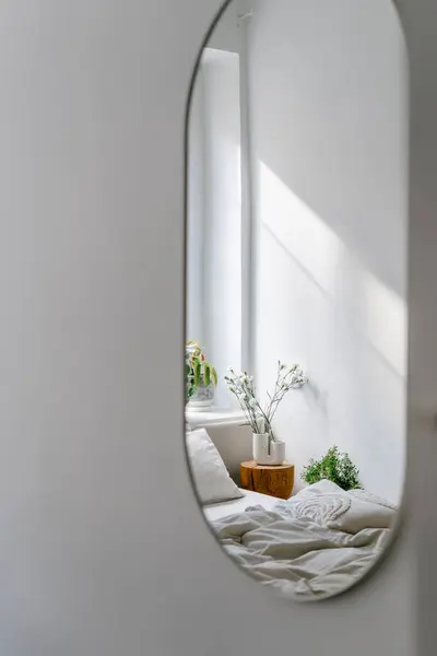 Vertical shot of oval mirror hanging on grey wall in bedroom. Reflection of bed with bedding and side table in room. Minimalist interior at home. Copy space.
