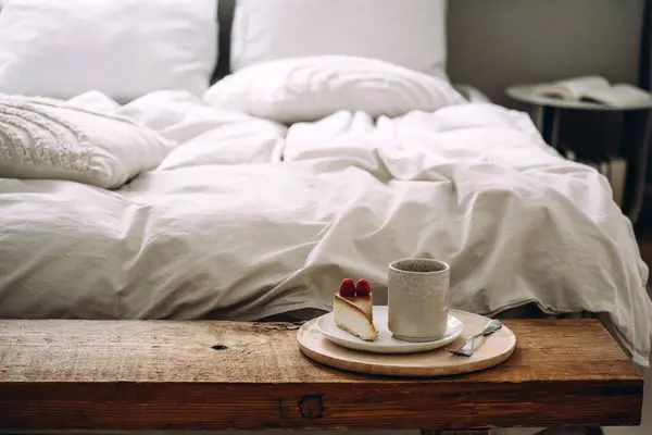 Selective focus on bamboo board with ceramic cup with hot beverage and plate with piece of cake on wooden bench near comfortable bed with linen bedding in cozy bedroom in apartment