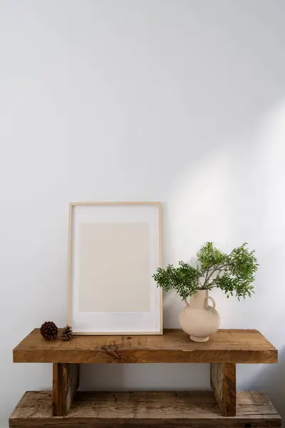 Stylish interior composition with wooden bench, mockup picture in frame and ceramic vase with green plant branch near white wall with copy space. Furniture from wood. Scandinavian style