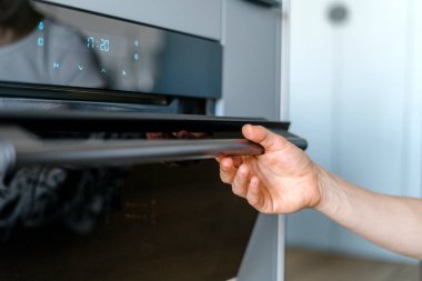 Modern mutfakta kadın el ele tutuşmaya ve elektrikli fırının cam kapağını açmaya odaklan. Sayısal zamanlayıcı ve fırın için ev aletinin düğmelerine sahip kontrol paneli.
