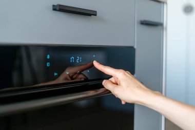 Yemek pişirmek için modern mutfak ekipmanlarının sensör tuşuna basan kadınlara odaklan. Kontrol paneli ve zamanlayıcısı olan elektrikli fırın. Bulanık gri mobilya arka planı.