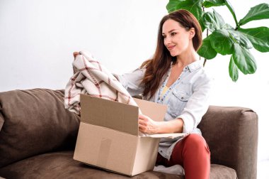 Evdeki koltukta oturan gülümseyen genç bir kadın, kutuları açıyor, internet siparişinden memnun. Ellerinde yumuşak ekose kumaş tutuyor. Beklenen kalite. Online mağaza servisinden memnun müşteri.
