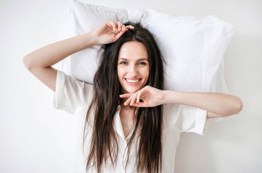 Mutlu ve neşeli bir kadının stüdyo portresi beyaz yastığın üzerinde uyanır, gülümser ve kameraya bakar. Kadın rahat bir uyku elbisesi içinde evde dinleniyor. Sağlıklı uykudan sonra günaydın.