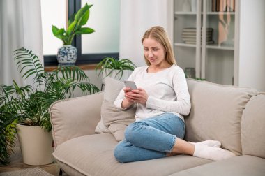 Bej koltukta oturan ve elinde akıllı telefon tutan gülümseyen kadın. İnternet sitesinde kadın arama, çevrimiçi alışveriş, internet üzerinden iletişim, mobil uygulama kurulumu