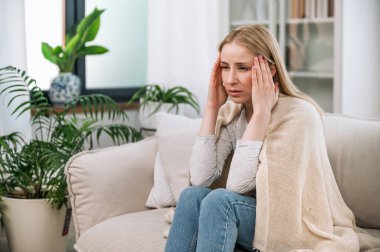 Kanepede oturan genç hasta bir kadın. Sıcak kareli kumaşlarla kaplı. Tapınakları tutuyor ve baş ağrısı ve ateş hissediyor. Migreni olan bir kadını üzmek ve evde kalmak. sağlık sorunu