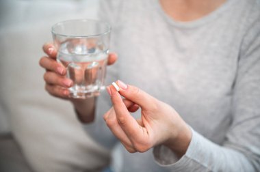 Kadın elindeki haplara özen gösteriyor. Grip belirtileri gösteriyor ve rahatlamak için ilaç alıyor. Bir bardak su ve ağrı kesici tabletle kesilmiş bir kadın resmi. sağlık hizmeti kavramı