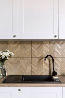 Beyaz dolaplı klasik mutfakta siyah lavabo ve musluk, ahşap tezgah, fayanslı duvar ve çiçek açan cam vazo. İç detaylar. Şık bir dairede yeni su musluğunun dikey çekimi