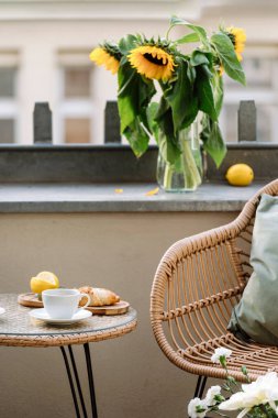 Yuvarlak masaya odaklan, üzerinde kahve ve kruvasan olsun. Yaz terasındaki Rattan sandalyesi cam vazoda ayçiçeklerinin yakınında. Balkonda dinlenme yeri. Binanın içinde bir avlu.