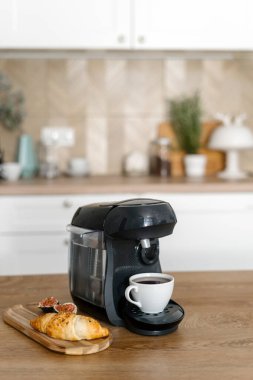 Tahta tabakta taze kruvasan ve bulanık mutfak arka planında, ahşap masadaki kahve makinesinde hazırlanmış bir fincan espresso üzerine seçici bir odak. Kafein içeceği ve pasta. Kahvaltı konsepti.