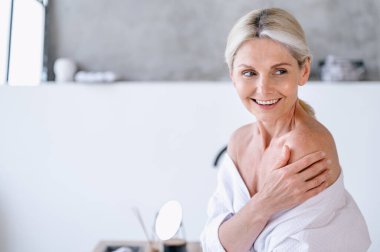 Bornozlu, çıplak omzuna dokunan, duştan sonra banyoda vakit geçiren mutlu yaşlı bir kadının portresi. Şımartma, güzellik ve spa konseptleri. Boşluk arkaplanı kopyala