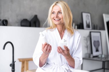 Cilt bakımı için iki farklı kozmetik şişesi tutan gülümseyen olgun kadın cilde ne uygulanacağını seçiyor. Orta yaşlı kadın bakım ürünlerini spa için karşılaştırıyor. Kişisel bakım, güzellik tedavisi