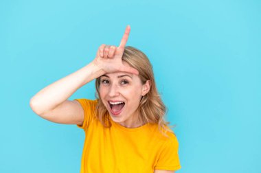 Young woman in yellow t-shirt showing loser gesture by fingers on forehead, teasing and mocking isolated on blue background. Female making fun of people and laughing clipart