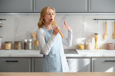 Mavi önlüklü kadın elinde tahta kaşıkla mikrofon gibi şarkı söylüyor, mutfakta dans ediyor ve kahvaltı hazırlıyor. İyi ruh hali kavramları. Ev hanımı müzik dinlerken eğleniyor, sağlıklı bir yemek hazırlıyor.