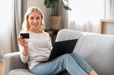 Evde dizüstü bilgisayarla rahat bir koltukta otururken kredi kartı gösteren bir kadın. Kolay bankacılık, para transfer sistemi, sanal cüzdan, müşteri hizmetleri ve İnternet üzerinden alışveriş kavramı