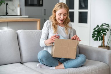 Aşırı neşeli genç bir kadın, karton kutuyu internetten gelen eşyalarla açıyor. Kadın acil teslimat paketini bekliyordu. Mutlu müşteri ya da memnun müşteri koltukta otururken internetten sipariş aldı.