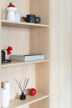 Ahşap kitaplıkta ya da dolapta açık alanda dergi, seramik çaydanlık, vazoda çiçek ve aroma çubukları var. Oturma odasında düzenli depo ve ahşap raflarda ev dekorasyonu.