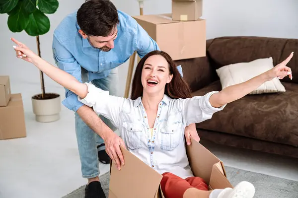 First time home buying, moving day concept. Happy young couple having fun, unpacking things in new flat, laughing. Woman sitting in cardboard box, man pushing her in house apartment