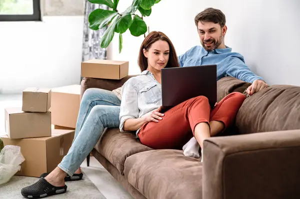 Young couple rent new apartment, prepare to relocation, sitting together on comfort couch near packed boxes and search moving transportation service in internet on laptop.