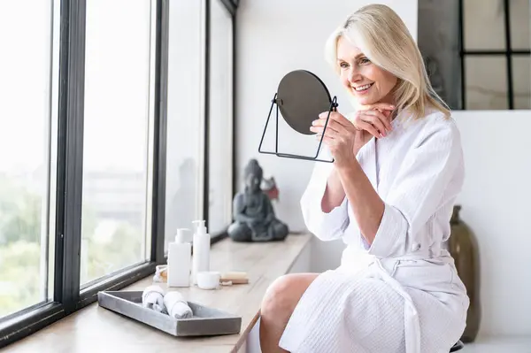 stock image Smiling mature woman looking at mirror and enjoy her healthy soft face skin. Female doing skincare rituals and using delicate cleanser and lifting cream. Self care concept