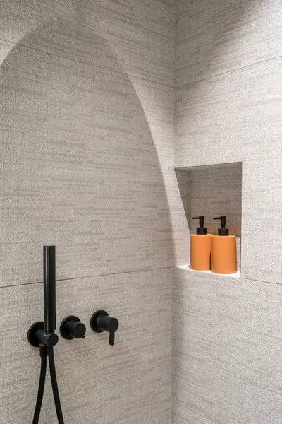 Detail in modern bathroom with minimalist style, black matte shower system with sprinkler on wall with textured tile and cosmetic products in dispenser bottles keeping in niche at luxury apartment