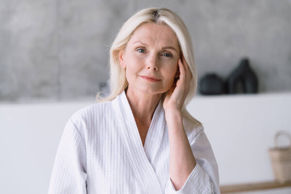 Adult senior woman in white bathrobe touching her perfect skin. Beautiful portrait of mid 50s aged female. Advertising anti age, rejuvenation, face lifting products, home spa and beauty treatment