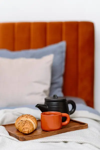 Selective focus on bamboo tray with ceramic teapot, cup and fresh croissant on comfortable bed with bedding. Tasty breakfast in apartment. Cozy home interior