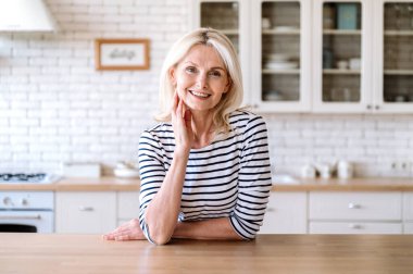 Neşeli orta yaşlı kadın mutfak masasının yanında duruyor, gülümsüyor ve kameraya bakıyor. Kendine güvenen neşeli kadın girişimci portresi. 