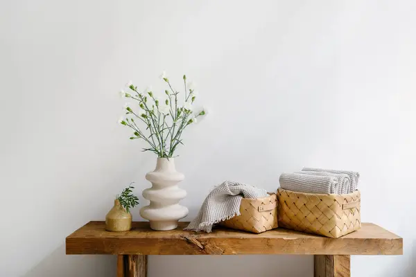 stock image Wooden bench with white flowers in clay vase close to wicker box with fresh, clean shower towels against bathroom wall on background. Concepts of personal accessory, hygiene and grooming products