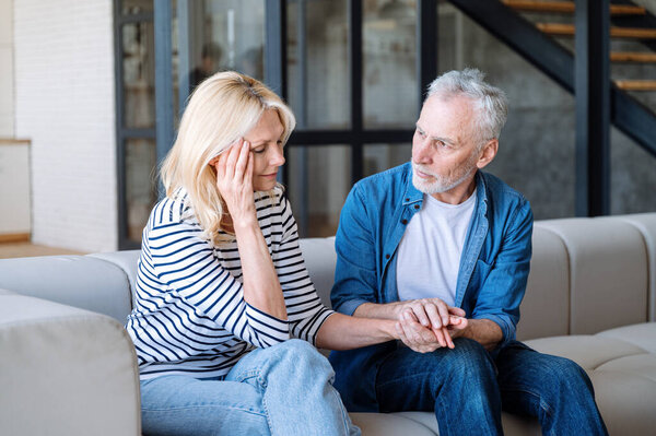 Husband comforting and showing support to his tired wife with headache. Concept of problems with memory and mental health disease. Caring man hold hand of upset woman while she sitting on sofa