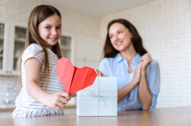 Çocuk elinde kalp şeklinde kırmızı el yapımı origami kartpostala odaklan. Annemin doğum günü hediyesi. Çocuk evde ebeveyn için hediye kutusu veriyor. Tatil, kutlama konsepti.