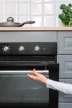 Paslanmaz çelik sapı olan kadının el kamerası açık ve mutfakta elektrikli fırın açıyor. Kadınlar modern ev aletleriyle evde yemek hazırlıyorlar. Pişirme kavramı.