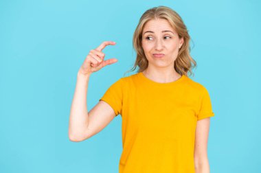 Young woman standing over blue background and showing small size gesture with hand. Sceptic female in yellow t-shirt doing sign little with fingers. clipart