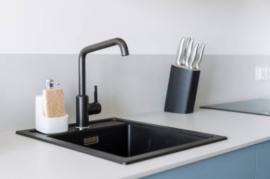Modern granit lavabo ve ev mutfağında siyah su musluğu. Yeni musluk ve bulaşık deterjanı şişesi. Dolabın tezgahında sünger var. Dairenin içinde minimalist iç mekan var.