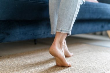 Kırpılmış çıplak ayaklı kadın bacakları mavi kanepenin yanında yürüyor. Evdeki rahat oturma odasında dokuma halıda dikilen kot pantolonlu genç kadının yakın görüntüsü.