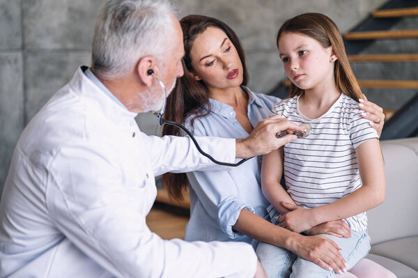 Врач-педиатр в медицинском халате проверяет пациентку, слушает дыхание при помощи стетоскопа во время домашнего визита. Грустный ребенок плохо себя чувствует и у него бронхит. Концепция профилактики пневмонии.