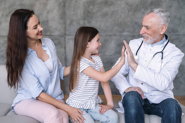Улыбающийся ребенок, сидящий на диване рядом с матерью, дает пять педиатру на домашней встрече. Пациентка счастлива после обследования семейным врачом, чувствует себя хорошо и выздоровел. Концепция здравоохранения