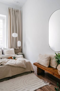 Rahat bir yatak odası, ahşap bir bank ve oval aynayla dikey çekimler. Modern odada bej renkli tekstil. Tarafsız renklerde ev dekoru.