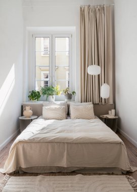 Çarşaflı, battaniyeli ve yastıklı rahat bir çift kişilik yatak vazolu gece masaları, kolye lambaları perdeli saksılar pencere perdeleri ve yatak odasının zemininde hafif iç döşemeli halı.