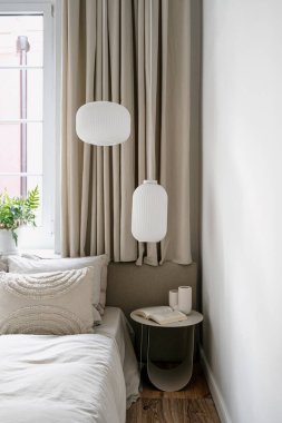 Yataklı rahat bir yatağın dikey görüntüsü, rahat bir evde klasik tarz yatak odasının penceresinin yanında kitap bulunan yan masa. Ahşap zeminin üstündeki odada tavan lambaları..