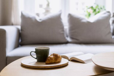 Tahta tabakta taze kruvasan ve seramik fincanda kahve ve yuvarlak masanın üzerinde açık bir kitap üzerine odaklanın. Kahvaltıyı evde yaparız. Oturma odasının içi rahat.