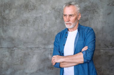 Gri arka planda izole edilmiş kendinden emin olgun bir adamın portresi. Sıradan giysiler içinde iş adamı yan yana bakıyor, kollarını kavuşturuyor. Liderlik kavramı.