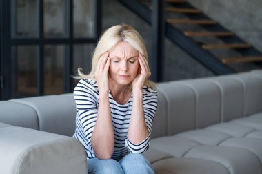 Akıl sağlığı sorunları konsepti. Orta yaşlı bir kadın kanepeye oturmuş, gözleri kapalı, şakaklarına dokunuyor, migreni var, başı ağrıyor. Yorgunluk ve yorgunluk.