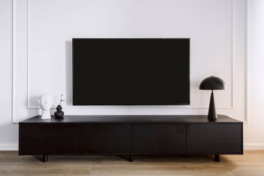 Oturma odasındaki beyaz duvarda asılmış, içinde minimalist iç mekan olan modern bir televizyon ekranı. Kara tahta aletin altında duruyor. Ev dekoru ve lamba dolapta. Ev elektroniği kavramı