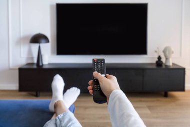 Oturma odasındaki konforlu koltukta dinlenirken kadın el kumandasına odaklan ve duvardaki televizyon panelini aç. POV 'lu kadın evde televizyon izlemek için kanal seçiyor