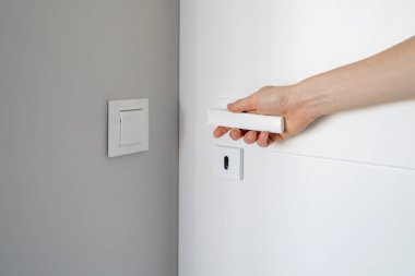 Kırpılmış kadın fotoğrafı daireye kapı tokmağını tutarken giriyor. Beyaz metal saplı, kadın eli açılış odasının kapısını kapatın. Modern dairede iç mekan elementi. Duvardaki ışık düğmesi