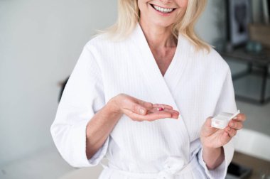 Bornozlu, gülümseyen orta yaşlı bir kadının avuç içinde multivitamin hapı tutarken, güzellik derisi ve saçı için vitamin tableti alırken çekilmiş bir fotoğrafı. Sağlıklı yaşam tarzı kavramı