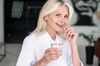 Gülümseyen orta yaşlı kadın bir bardak suyla sabahları güzellik ve sağlık için vitamin takviyesi hapı alıyor. Refah için multivitaminler ve mineraller.