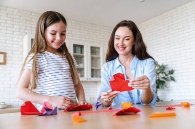 Gülümseyen anne ve kız kalp şeklinde kırmızı kağıtlardan kalpler yaratıyor, yemek odasında masada oturuyor. Hobi aktivitesi için boş zaman. Evde çocukla oynamak..