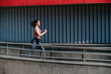 Spor giyimli genç bir sporcunun profiline göre şehirde hızla koşuyor. Sportif kadın koşuyor, fotokopi alanı olan şehir duvarının yakınına gidiyor. Kardiyo eğitimi alan bir kız. Egzersiz konsepti